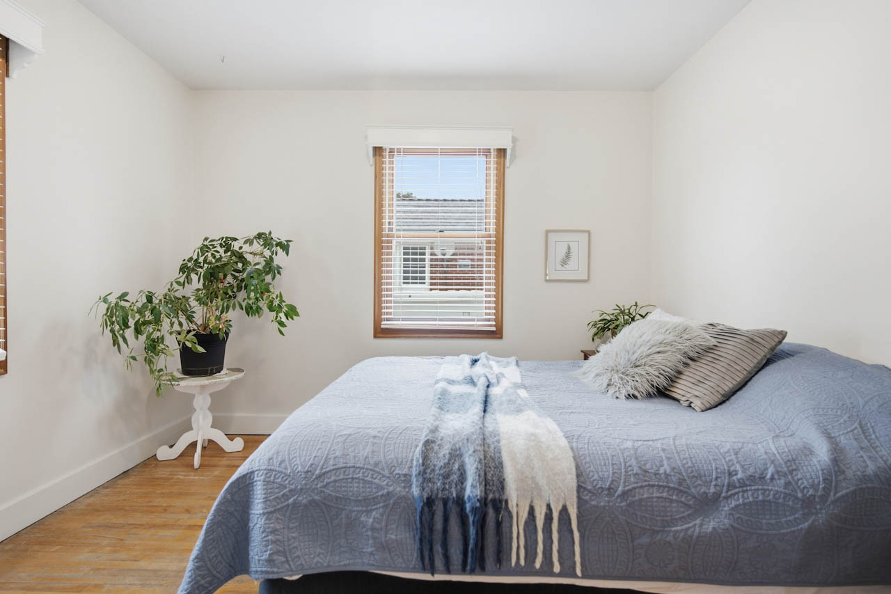 Bedroom with hardwood floor, white walls, wood-trimmed window, and a plant stand
