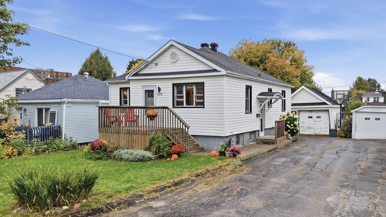 Angled view of the bungalow, front deck, and paved driveway leading to the detached garage